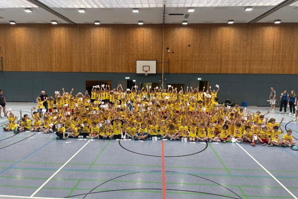 Gruppenszene von Kindern in gelben T-Shirts, die im Sporthalleninneren stehen und mit erhobenen Händen Posieren.