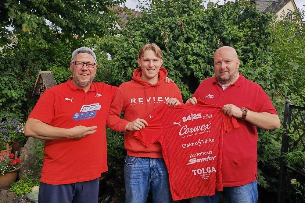 Drei Männer in roten T-Shirts posieren im Freien mit einem Trikot vor einem Garten.