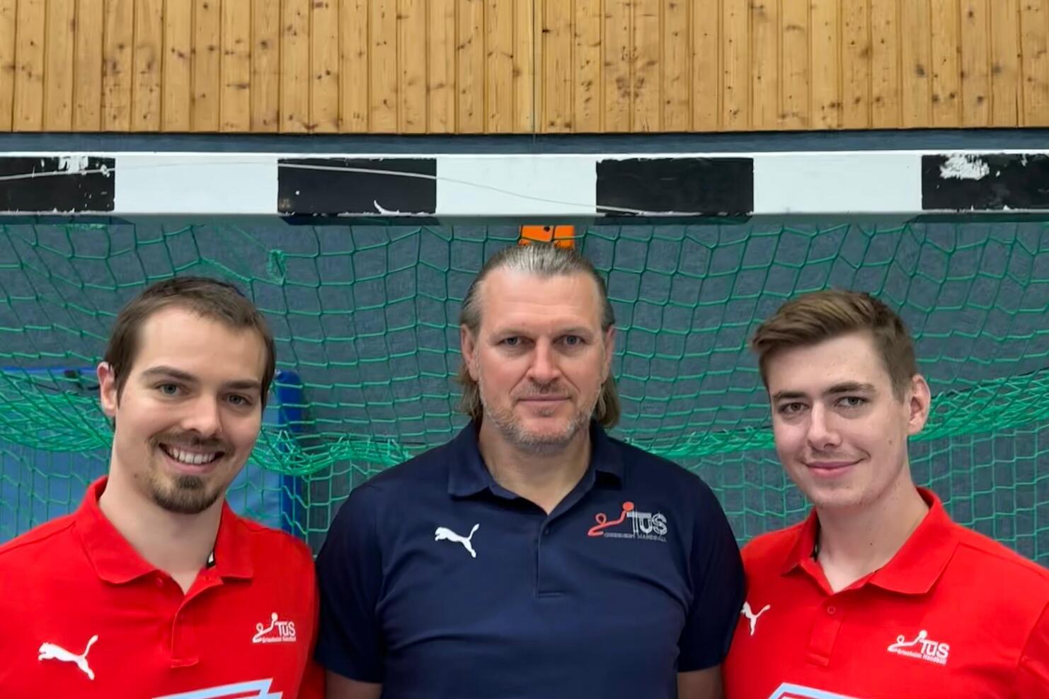 Drei Personen stehen vor einem grünen Handballtor, tragen sportliche Kleidung und lächeln in die Kamera.