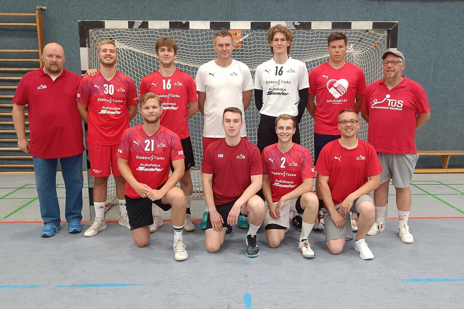 Mannschaftsbild einer Handballmannschaft in roter und schwarzer Sportkleidung vor einem Handballtor.