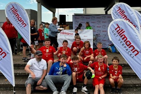 Gruppe von Kindern in roten Sporttrikots mit Medaillen sitzt auf Stufen, umgeben von Fahnen und Zuschauern.