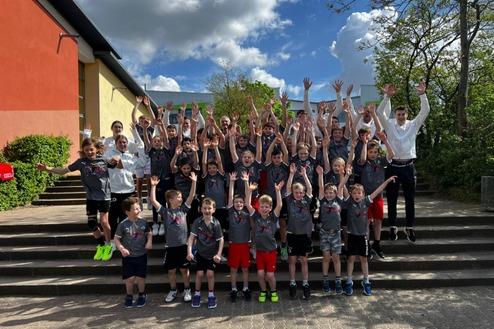 Gruppenshot von Kindern und Betreuern, die auf Treppen stehen und jubelnd die Hände heben, unter blauem Himmel.