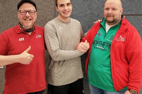 Drei Männer stehen in einer Sporthalle, einer schüttelt die Hand des anderen, während der Dritte Daumen hoch zeigt.