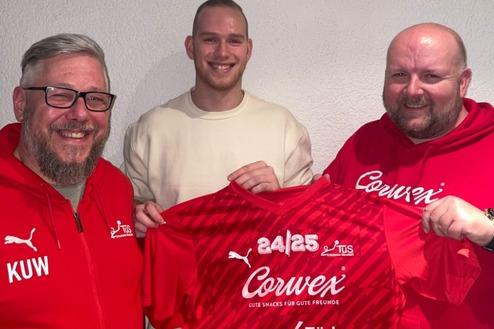 Drei Männer halten ein rotes Fußballtrikot mit Sponsorenlogos vor einer hellen Wand.