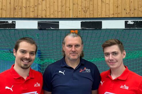 Drei Personen stehen vor einem grünen Handballtor, tragen sportliche Kleidung und lächeln in die Kamera.