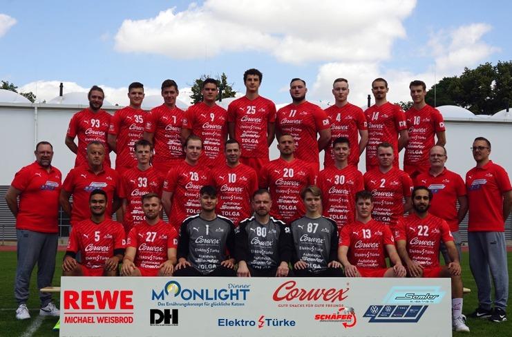 Mannschaftsfoto einer Fußballmannschaft in roten Trikots auf einem Sportplatz, stehend und sitzend vor einem Sponsorenschild.