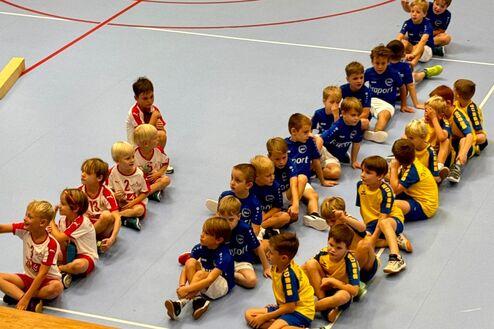 Gruppe von Kindern in Sportkleidung sitzt auf dem Boden einer Halle, einige lachen und unterhalten sich.