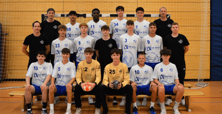 Gruppenbild einer Handballmannschaft mit Trainern, in einheitlichen Trikots und Shorts, vor einem Handballtor.