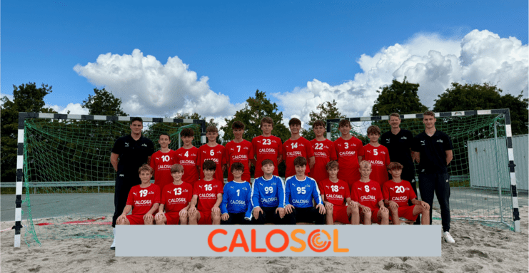 Gruppenfoto einer Jugendfußballmannschaft in roten Trikots vor einem Tor auf Sandplatz, begleitet von Trainern.