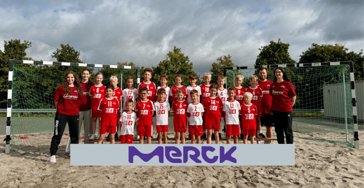 Eine Gruppe von Kindern und Trainern steht lächelnd vor einem Handballtor, eingekleidet in rote und weiße Sporttrikots.