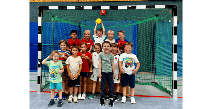 Gruppe von 14 Kindern steht vor einem Handballtor, ein Kind hält einen Handball hoch. Alle lächeln fröhlich.