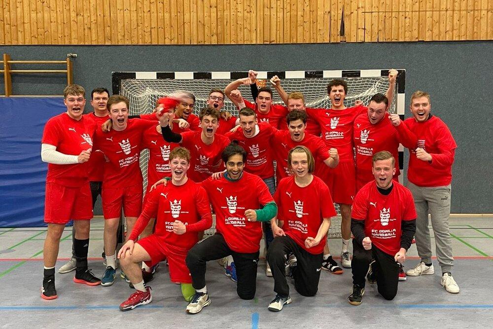Gruppe von 14 jungen Spielern in roten Trikots, jubelnd vor einem Handballtor in einer Sporthalle.
