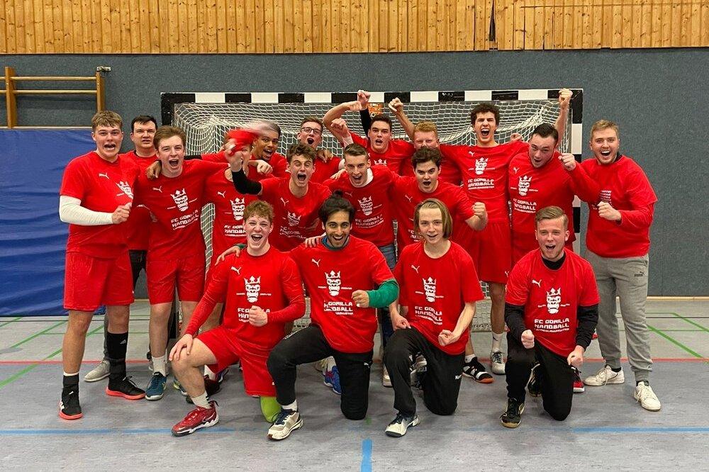 Gruppe von 14 jungen Spielern in roten Trikots, jubelnd vor einem Handballtor in einer Sporthalle.
