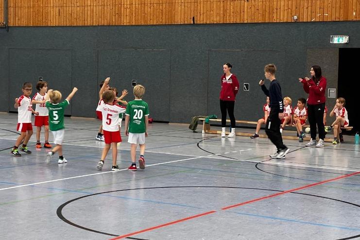 Kinder beim Handballspielen in Sportkleidung, Trainer beobachten das Spiel am Seitenrand.