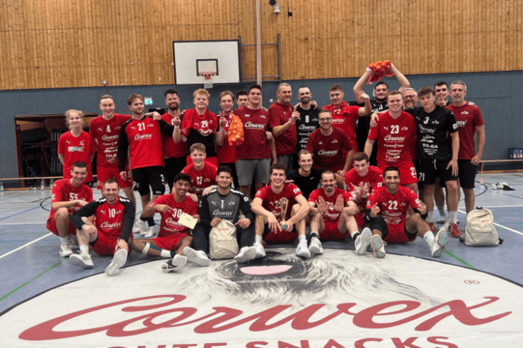 Gruppendfoto von 26 Handballspielern in roten Trikots auf einem Hallenboden mit Markenlogo und Slogan im Vordergrund.