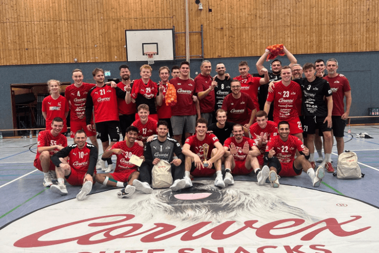 Gruppendfoto von 26 Handballspielern in roten Trikots auf einem Hallenboden mit Markenlogo und Slogan im Vordergrund.