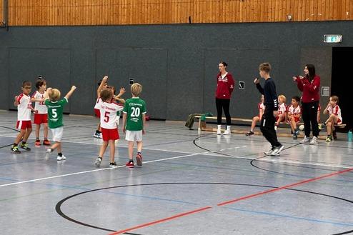 Kinder beim Handballspielen in Sportkleidung, Trainer beobachten das Spiel am Seitenrand.