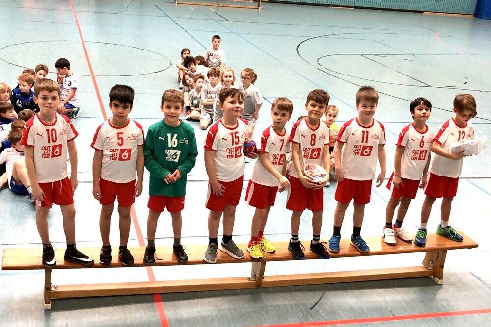 Acht Jungen in Sportkleidung stehen auf einer Bank in einer Sporthalle, im Hintergrund sitzen weitere Kinder.