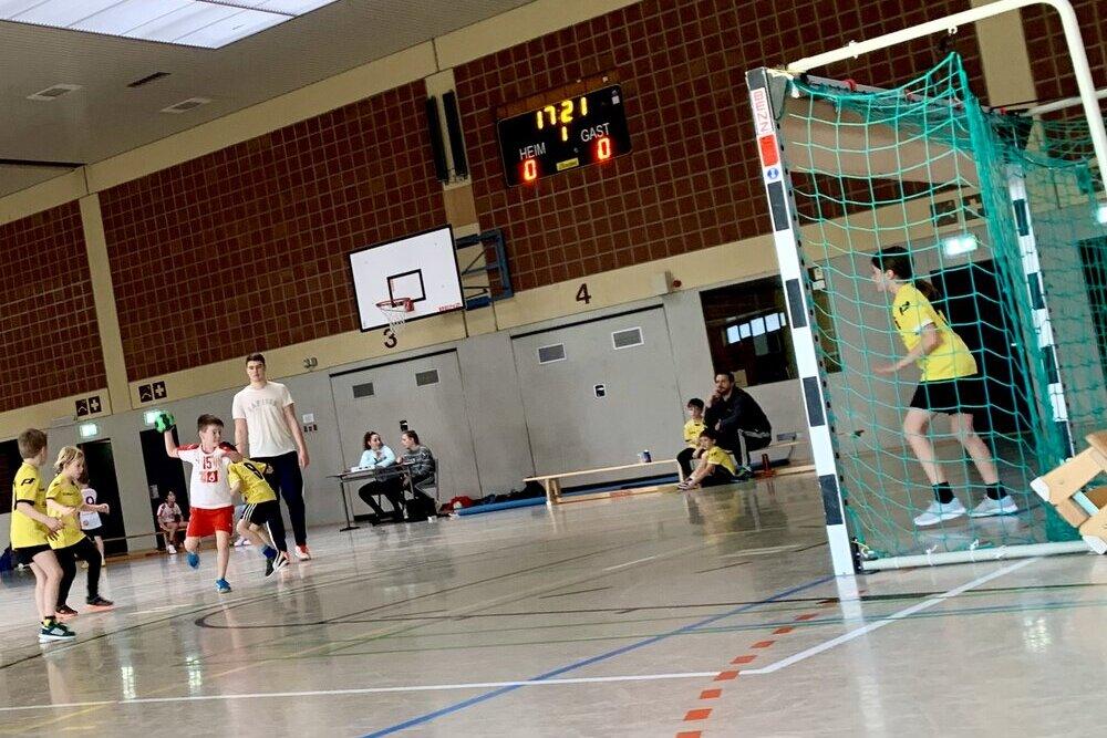 Kinder spielen Handball in einer Sporthalle, während ein Spieler einen Ball wirft. Die Anzeigetafel zeigt 0:0.