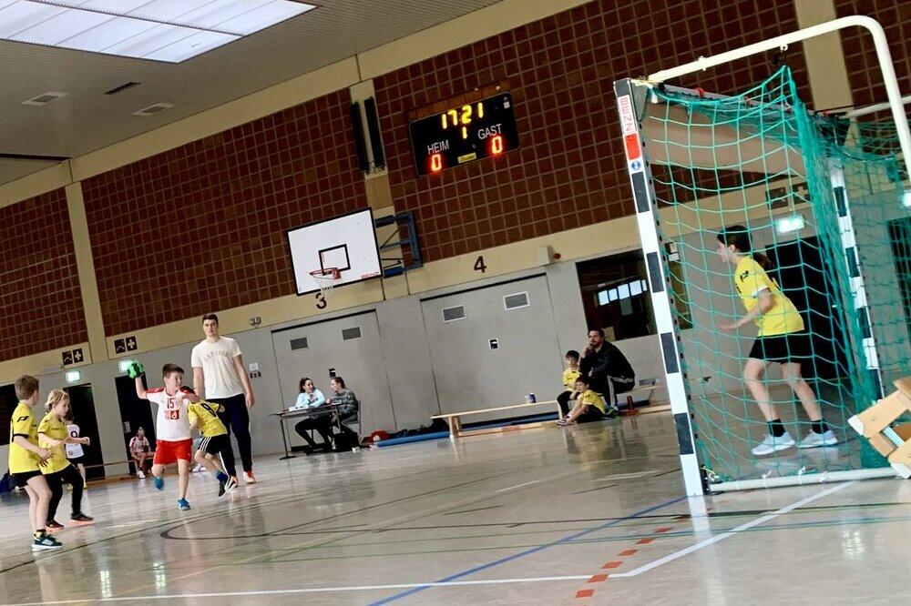 Kinder spielen Handball in einer Sporthalle, während ein Spieler einen Ball wirft. Die Anzeigetafel zeigt 0:0.