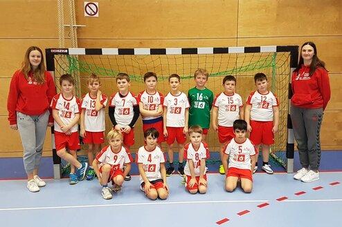 Gruppe von neun Kindern in Sportkleidung sowie zwei Trainerinnen stehen vor einem Handballtor in einer Sporthalle.