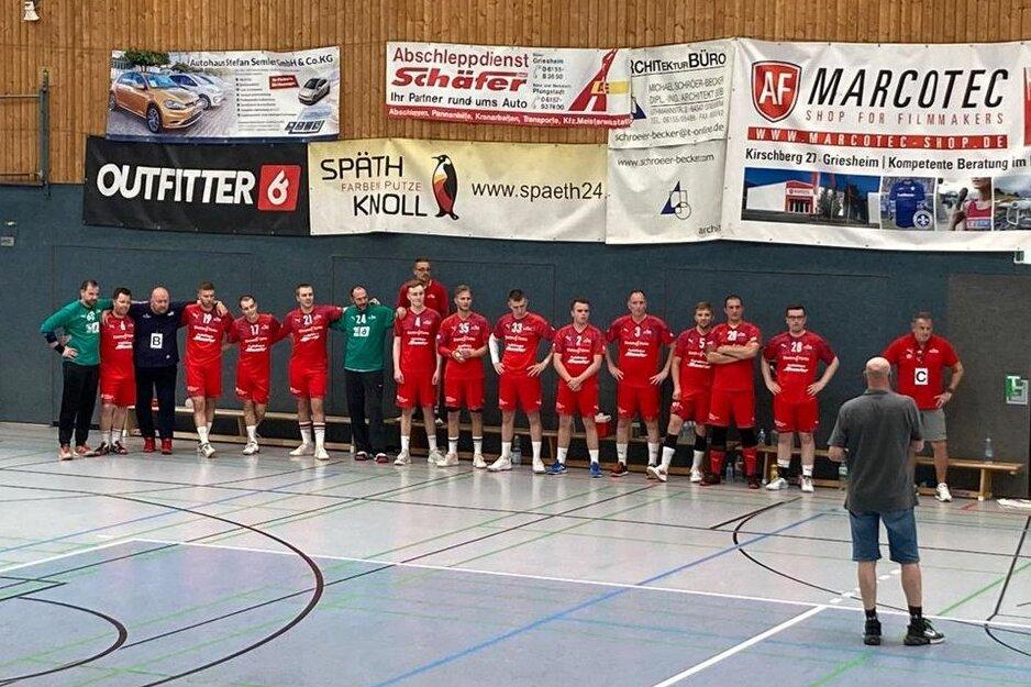 Mannschaft aus Männern in roten Trikots steht an der Wand einer Sporthalle, während ein Trainer sie ansprechen.