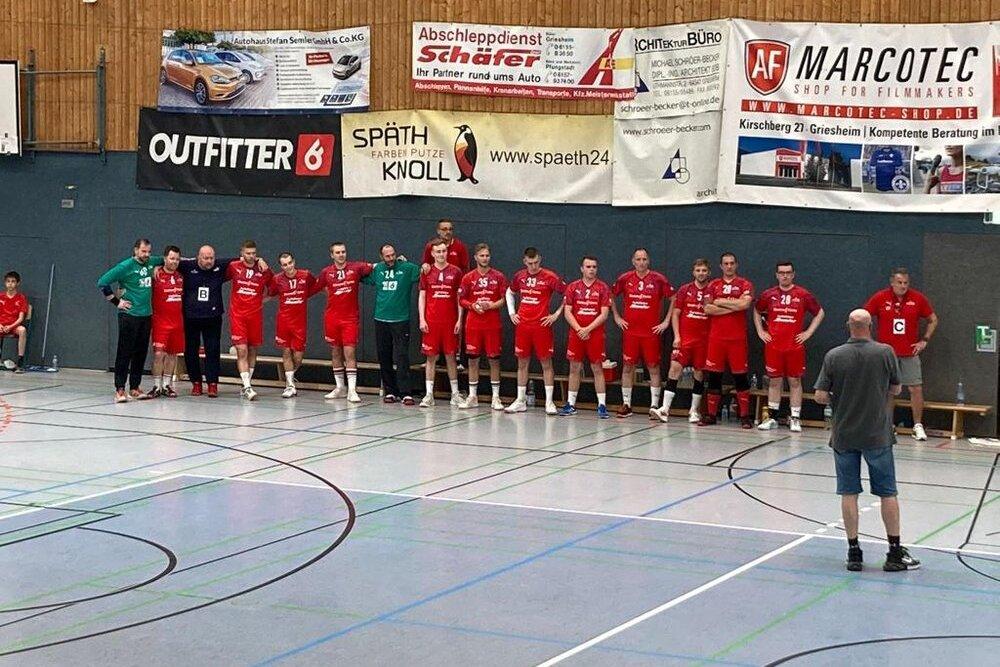 Mannschaft aus Männern in roten Trikots steht an der Wand einer Sporthalle, während ein Trainer sie ansprechen.