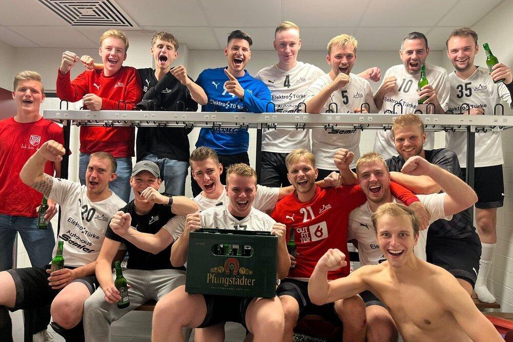 Gruppenfoto von Fußballspielern in einem Umkleideraum, feiernd mit Bierflaschen und einem Kasten auf einer Bank.