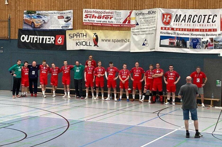 Mannschaft aus Männern in roten Trikots steht an der Wand einer Sporthalle, während ein Trainer sie ansprechen.