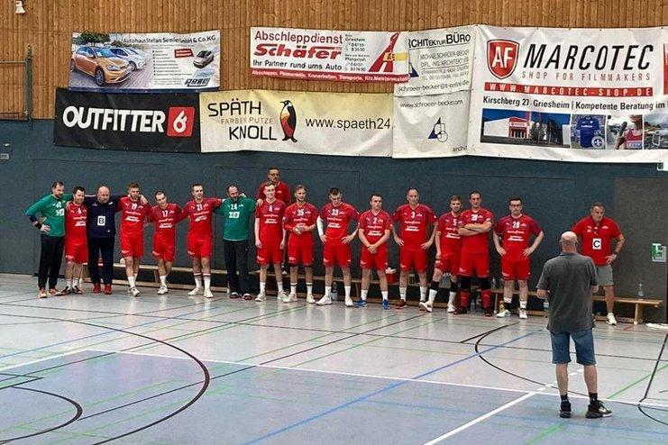 Mannschaft aus Männern in roten Trikots steht an der Wand einer Sporthalle, während ein Trainer sie ansprechen.