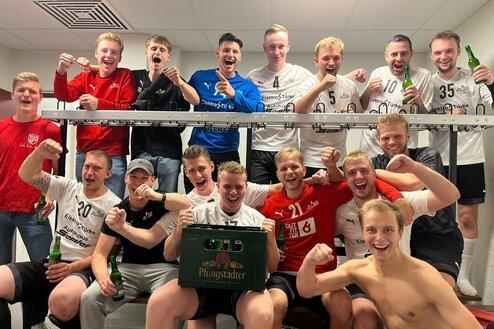 Gruppenfoto von Fu&szlig;ballspielern in einem Umkleideraum, feiernd mit Bierflaschen und einem Kasten auf einer Bank.