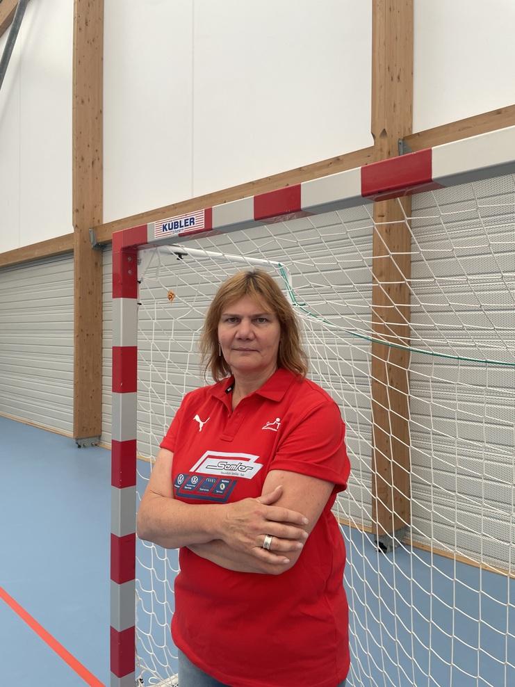 Frau mit rotem Polo-Shirt steht vor einem Handballtor in einer Sporthalle. Sie hat die Arme gekreuzt und schaut direkt.