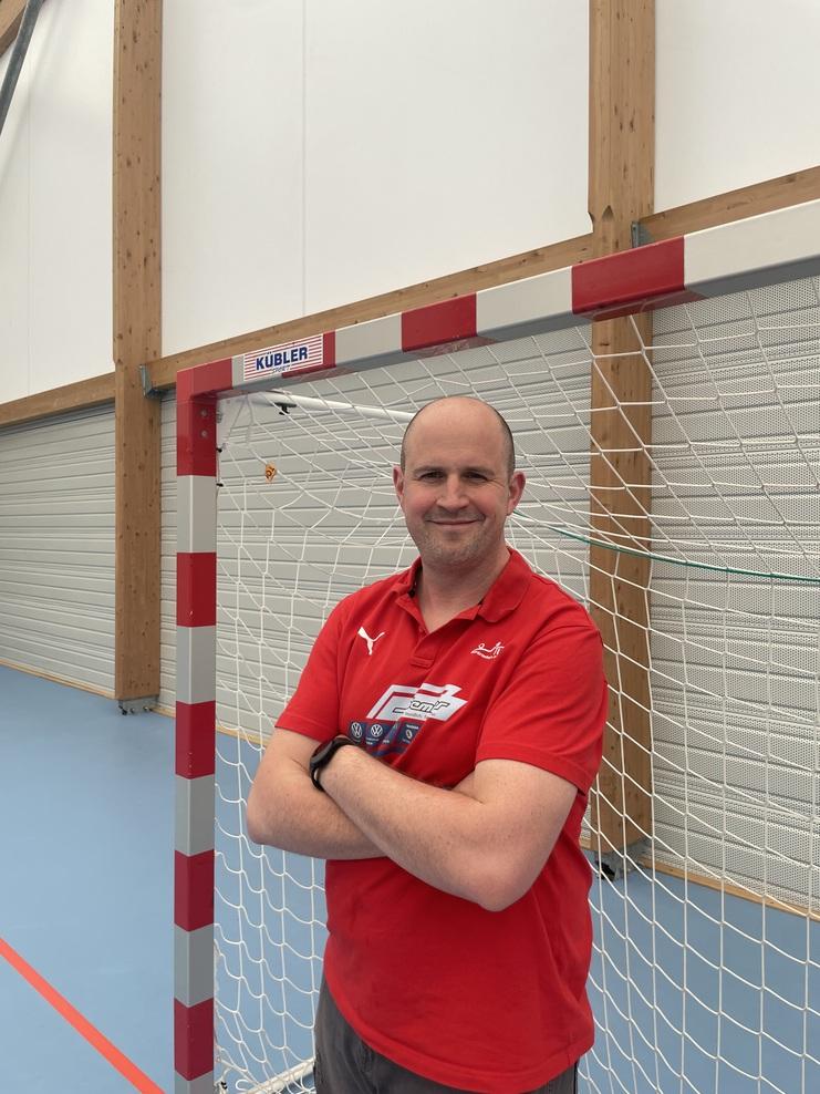 Mann mit verschränkten Armen in rotem Shirt steht vor einem Handballtor in einer Sporthalle.