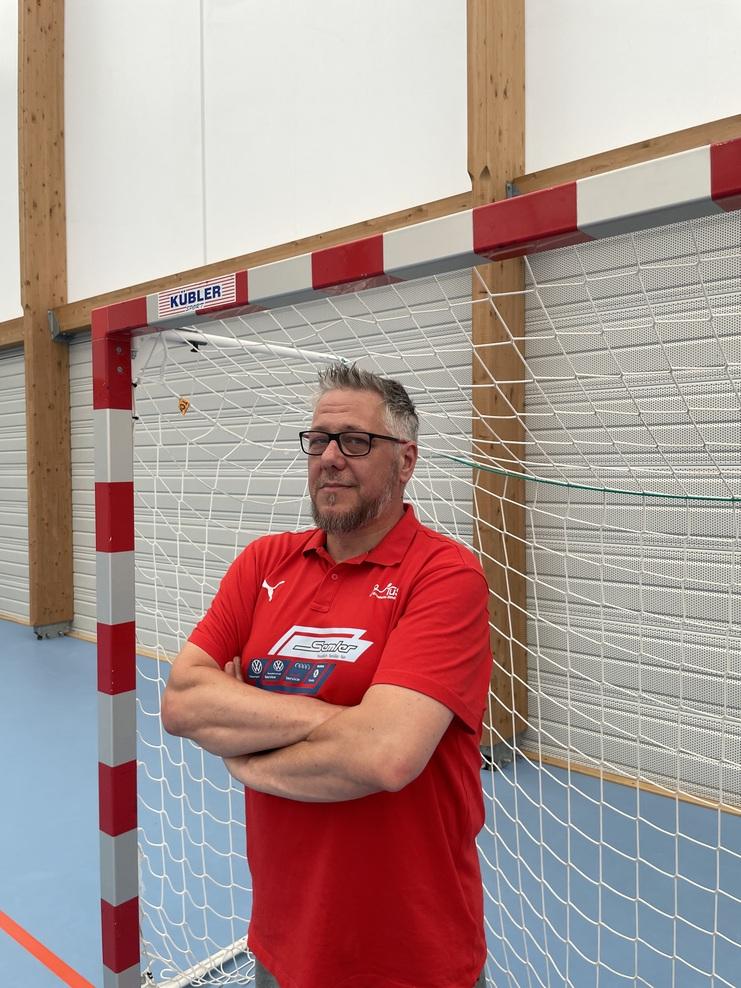 Mann in rotem Polohemd steht lächelnd vor einem Handballtor in einer Sporthalle.