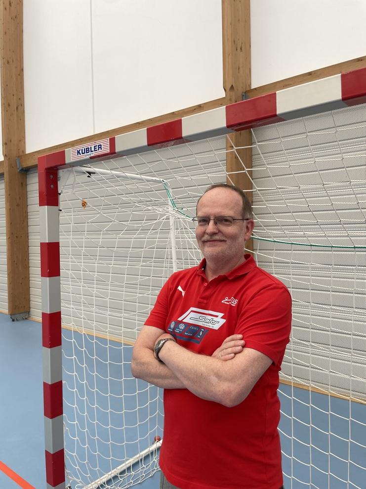 Mann mit Brille und rotem Shirt steht vor einem Handballtor in einer Sporthalle.