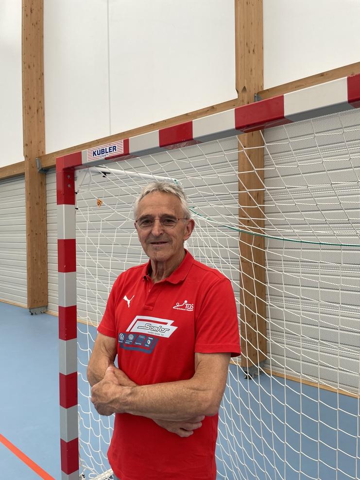 Älterer Mann in rotem Polo-Shirt steht neben einem Handballtor in einer Sporthalle. Holzbalken sind im Hintergrund sichtbar.