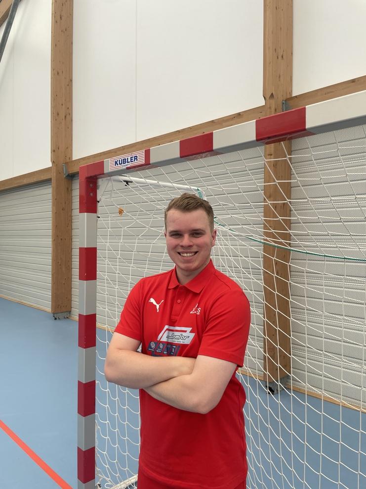 Junger Mann in rotem Polo, lächelnd vor einem Handballtor in einer Sporthalle, mit verschränkten Armen.