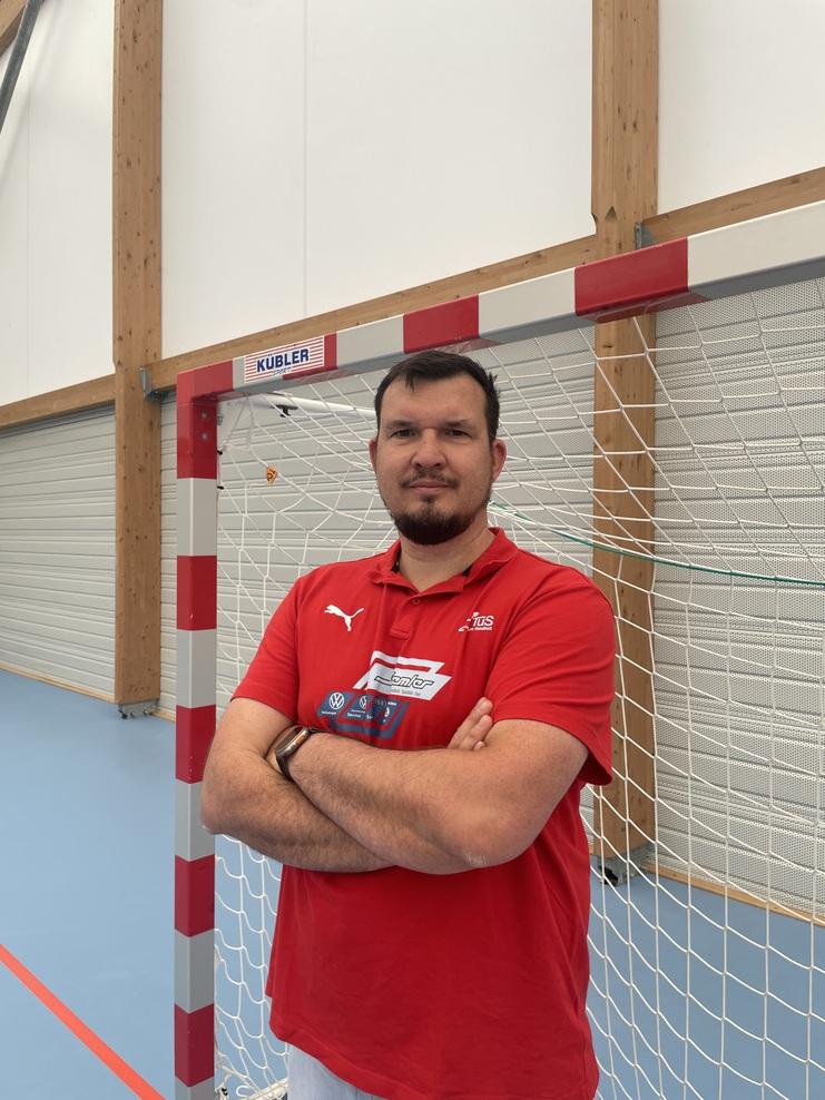 Mann mit verschränkten Armen steht vor einem Handballtor in einer Sporthalle. Er trägt ein rotes Sportshirt.