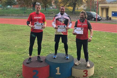 Drei Männer auf Siegerpodest in Sportbekleidung halten Urkunden, mit einer Laufbahn im Hintergrund und herbstlichen Bäumen.
