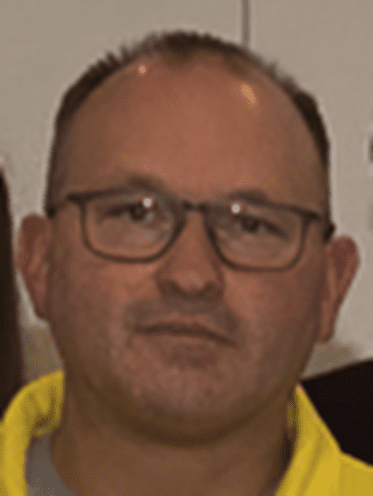 Nahaufnahme eines Mannes mit Brille und kurzen, dunklen Haaren, der ein gelbes Oberteil trägt. Hintergrund unscharf.