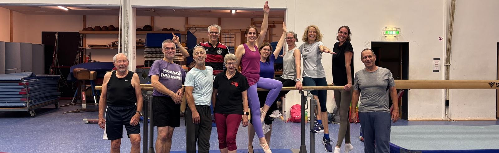 Gruppe von Sportlern in einer Turnhalle, l&auml;chelnd auf und vor einem Barren. Turnger&auml;te im Hintergrund sichtbar.