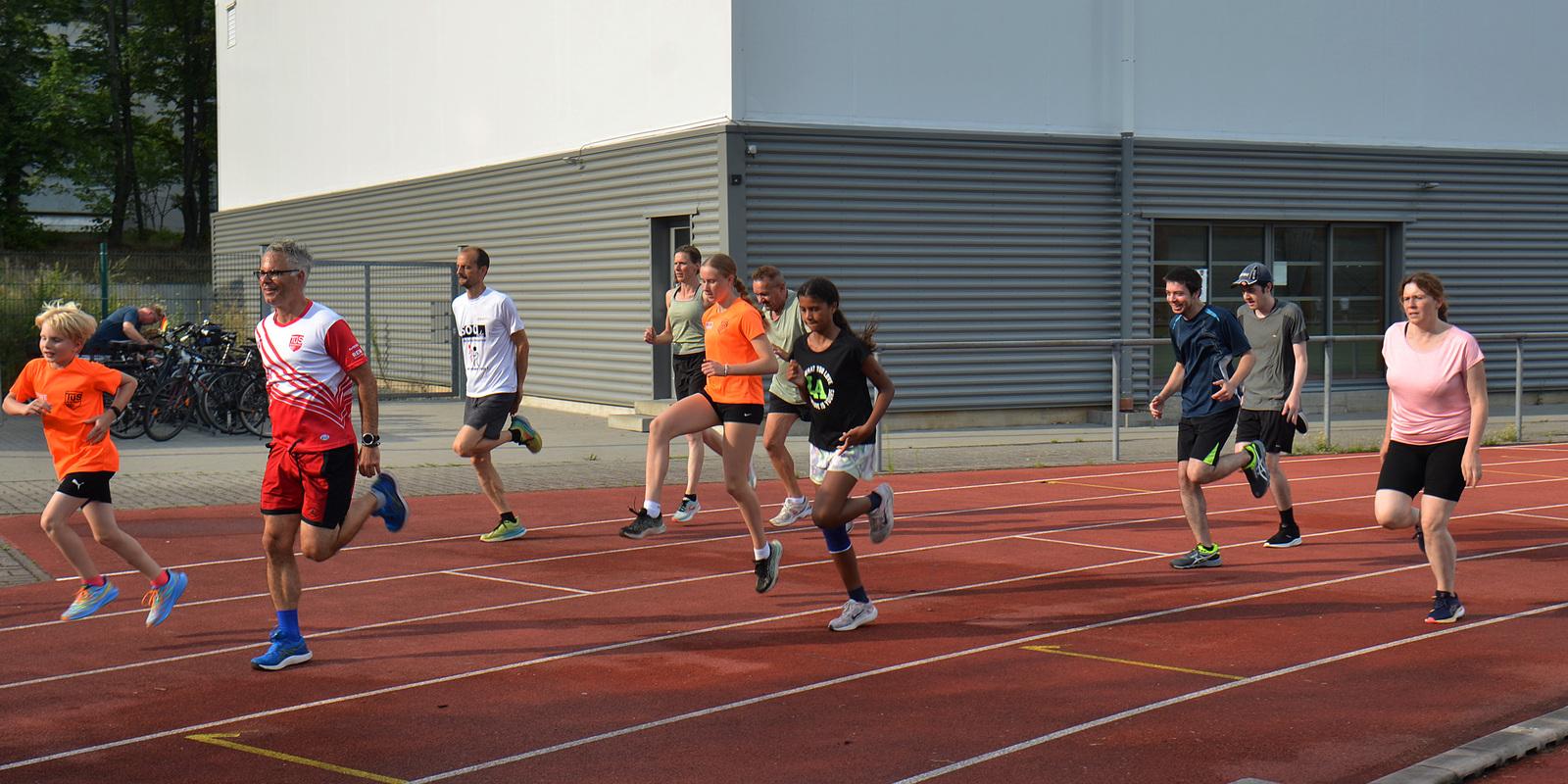 Gruppe von sechs L&auml;ufern in Sportkleidung, die auf einer roten Laufbahn joggen, im Hintergrund ein Geb&auml;ude.