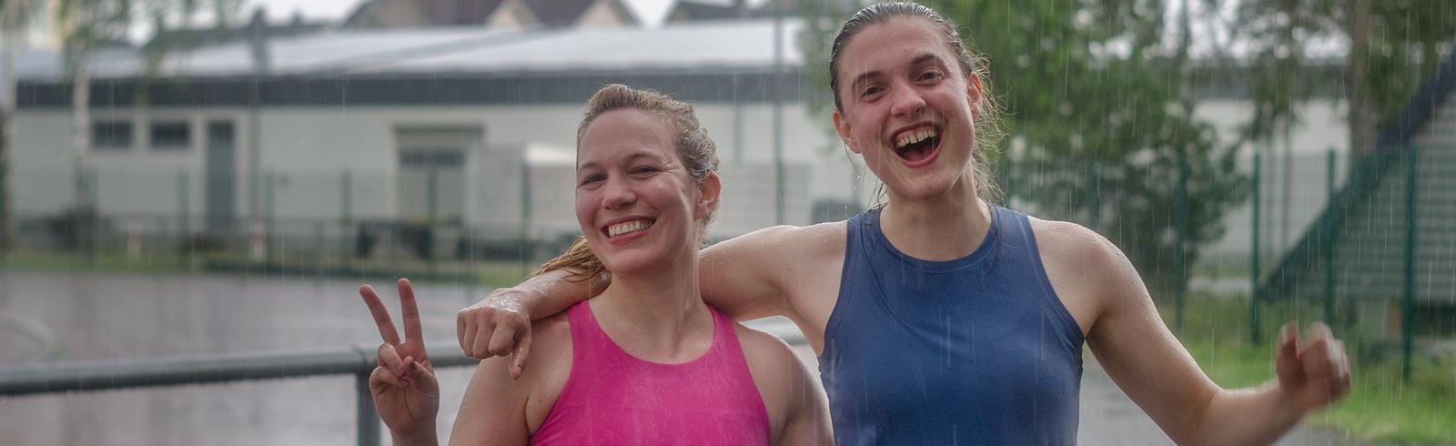 Zwei l&auml;chelnde Frauen posieren im Regen, eine zeigt ein Peace-Zeichen, im Hintergrund ein Sportplatz.