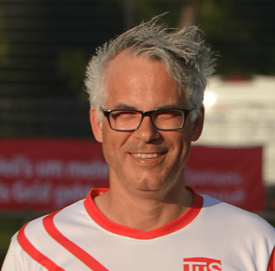 L&auml;chelnder Mann mit grauen Haaren und Brille, tr&auml;gt ein wei&szlig;es Trikot mit roten Akzenten, Hintergrund unscharf.