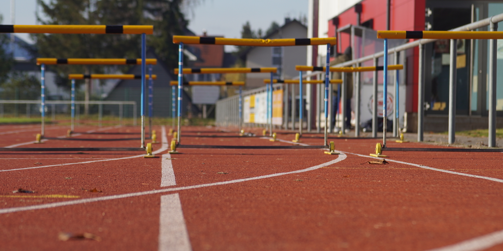 Blick auf eine Leichtathletikbahn mit H&uuml;rden in der N&auml;he und einem Sportgeb&auml;ude im Hintergrund.