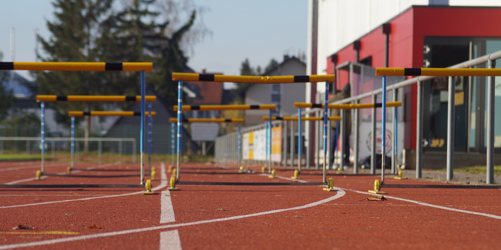 Blick auf eine Leichtathletikbahn mit H&uuml;rden in der N&auml;he und einem Sportgeb&auml;ude im Hintergrund.