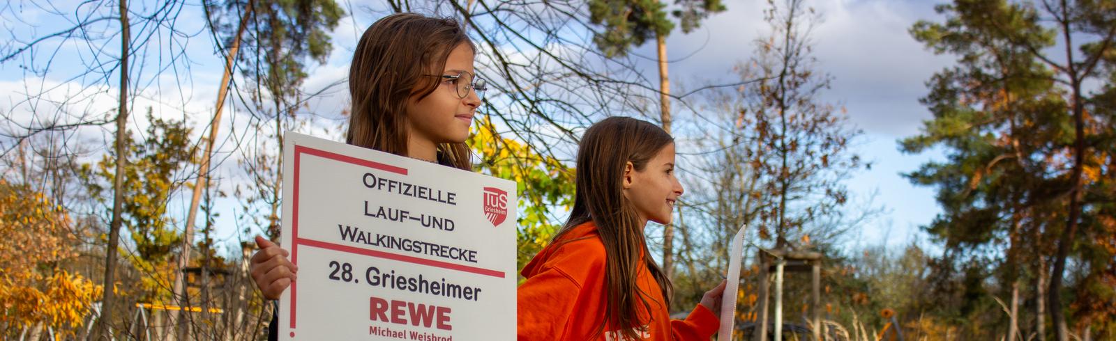 Zwei Mädchen halten Schilder für den Griesheimer Silvesterlauf in einem herbstlichen Park.