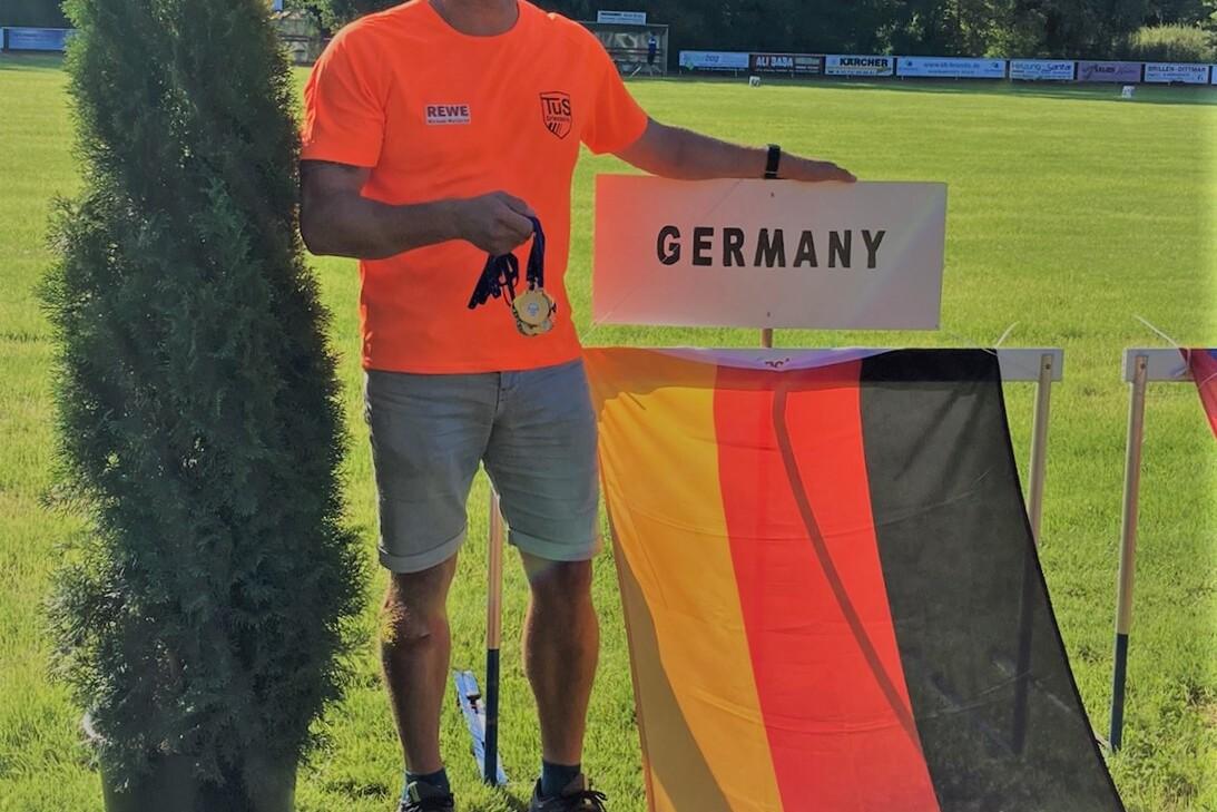 Mann in orangefarbenem Shirt hält eine Medaille vor einem Schild mit der Aufschrift „GERMANY“ und einer Deutschlandflagge.