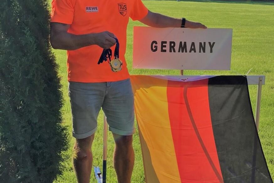 Mann in orangefarbenem Shirt hält eine Medaille vor einem Schild mit der Aufschrift „GERMANY“ und einer Deutschlandflagge.