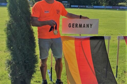 Mann in orangefarbenem Shirt hält eine Medaille vor einem Schild mit der Aufschrift „GERMANY“ und einer Deutschlandflagge.
